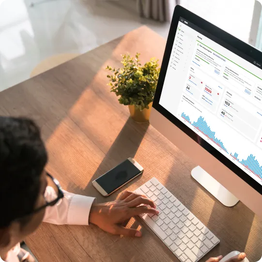 A security expert views the Duo admin dashboard on a computer, with a keyboard, mouse, smartphone, and plant on the desk. The dashboard shows various metrics and graphs.