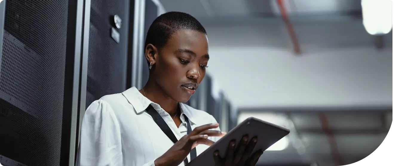 A woman in a white blouse stands in a server room, using a tablet while wearing an ID badge on a lanyard.