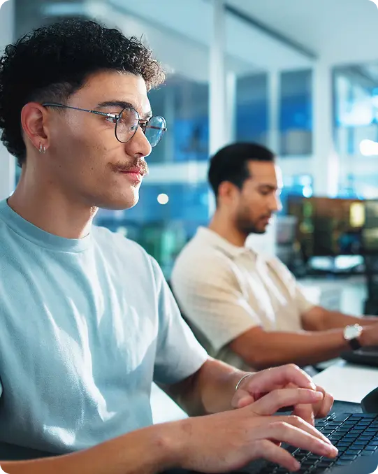 Two men type at computers in a bright, modern office space, one in focus and one in the background.