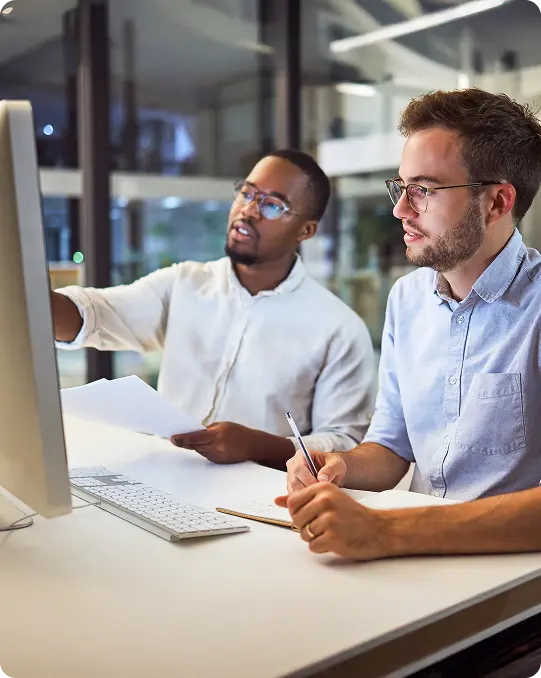Two colleagues in an office work at a computer, one pointing at the monitor while the other takes notes with a pen and notebook.