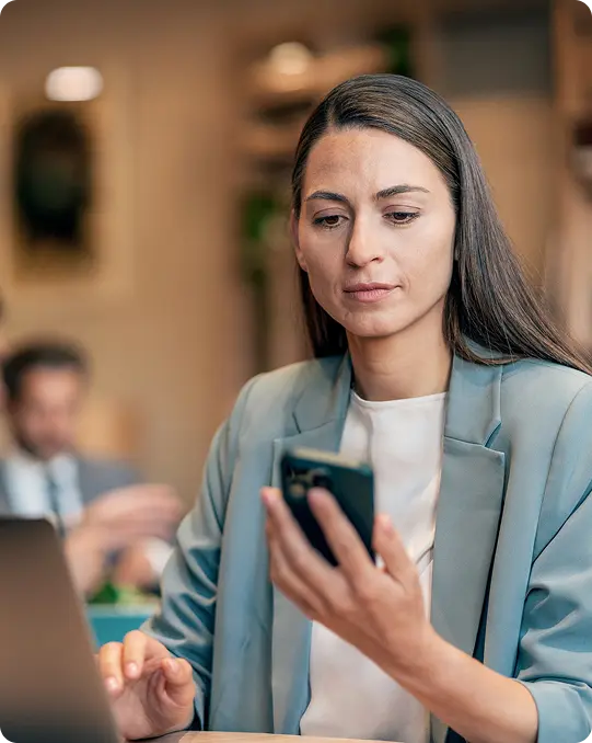 Woman in blazer sits at a desk with laptop, looking at her phone while two people talk in the background.