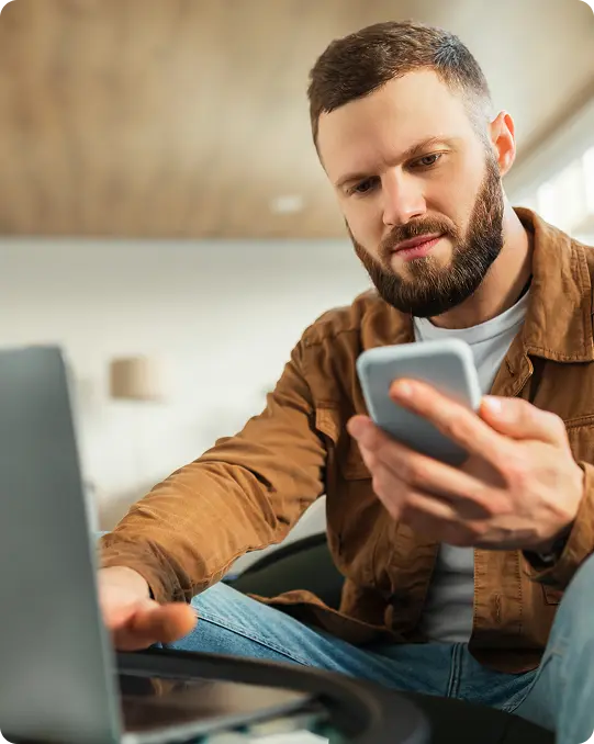 Man sitting at a table using a laptop while holding a smartphone, appearing focused as he works in a home or office setting
