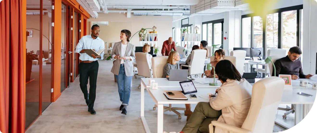 Open office with employees working at desks and two colleagues walking and talking in the aisle near glass-walled meeting rooms
