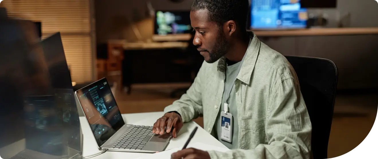 A man wearing a light green shirt and an ID badge works at a desk with a laptop, writing notes on a clipboard. Computer screens with data are visible in the background