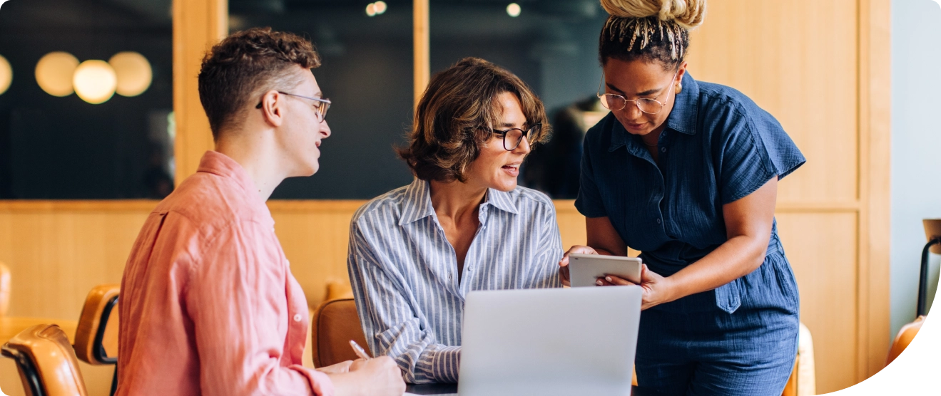 Three colleagues collaborate in an office, discussing something on a tablet and laptop
