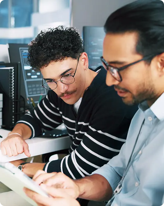 Two colleagues review information on a tablet in front of multiple computer monitors displaying code and technical data.