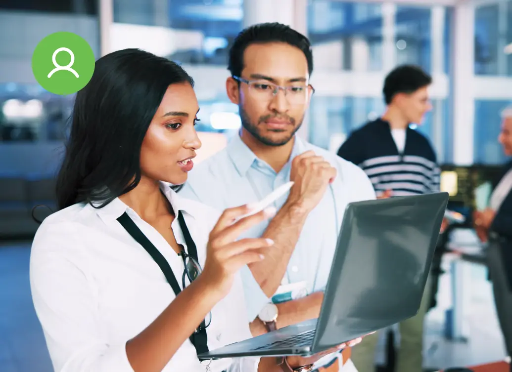 Two colleagues in an office review information on a laptop, while two others have a discussion in the background near workstations.