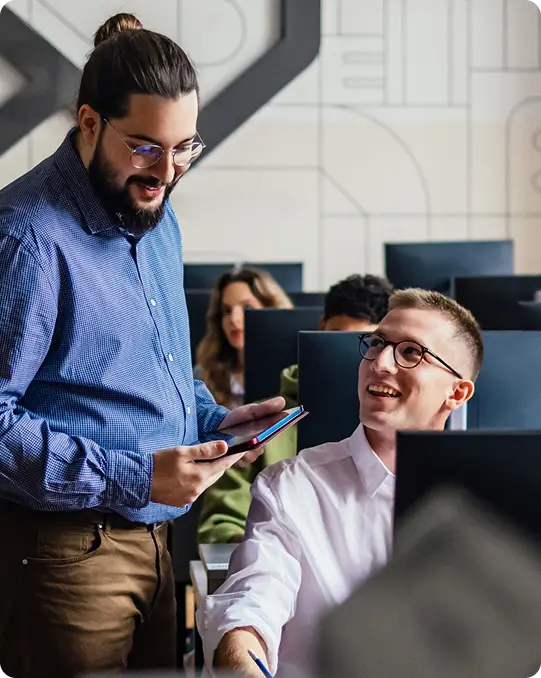 In an office, a man standing with a tablet speaks to a seated colleague who is smiling, while other people work at computers in the background.