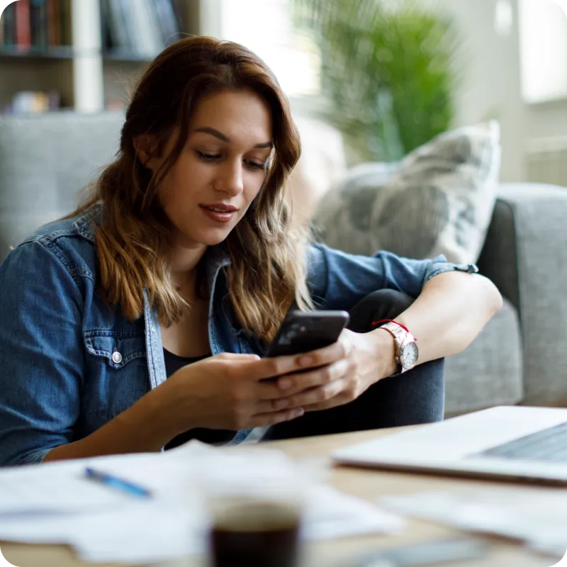 Person in a denim shirt sitting on a couch, holding a smartphone, with papers and a laptop on the table in front.