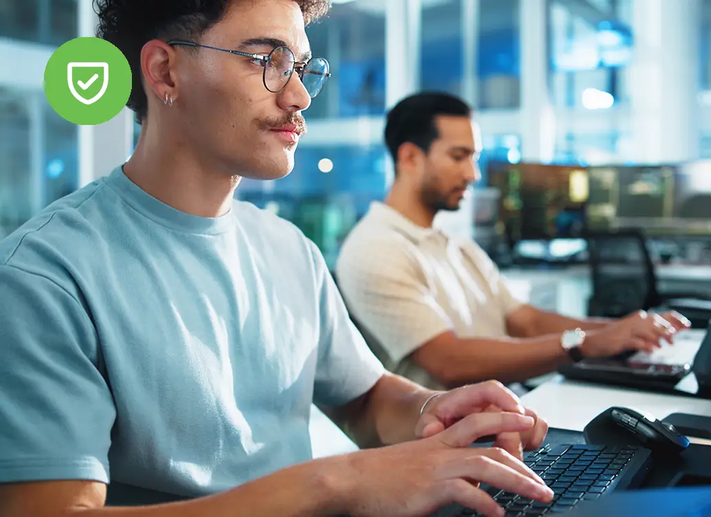 Two men type at computers in a bright, modern office space, one in focus and one in the background.