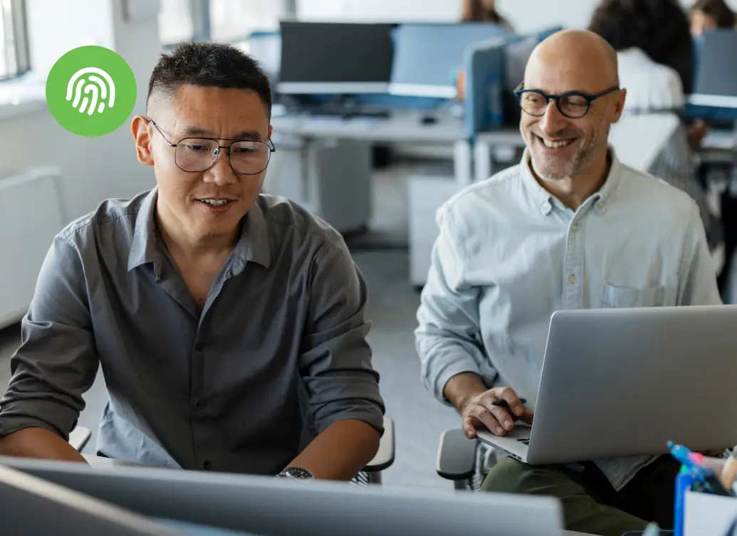 Two colleagues smiling while working at their computers in a bright office, with other employees focused at desks in the background